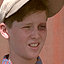 A pixelated close-up of a person wearing a dark cap with light trim, showing partial ear and mouth area against a pinkish background.