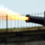 A cannon fires a bright yellow-orange muzzle flash against a light blue sky with faint clouds, positioned on a grassy terrain with a fence in the background.