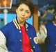 A person in a blue and white varsity jacket with a red tie stands against a blurred background of people and lights, with subtle changes in facial expression and head position across frames.