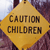 Yellow diamond-shaped sign with bold black text reading 'CAUTION CHILDREN' on a blurred background of purple and blue tones.