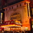 A pixelated static image of a theater interior with warm orange and brown tones, showing a stage, seating, and architectural details.
