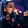 A person with long dark hair in a high ponytail sings into a microphone, wearing a black top with pink accents and a choker necklace, with red text appearing sequentially below.