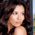 Pixelated close-up of a person with long dark brown hair, light skin, and a neutral expression against a blurred background.