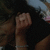 A person with long dark hair and a black top holds a small object near their face, wearing a silver bracelet on the wrist.