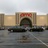 Static image of a building facade with a central dark arched entrance flanked by light-colored columns under a red horizontal band.
