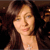 A pixelated close-up of a person with long dark hair, light skin, and a neutral expression against a dark background.