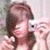 A person with brown hair holds a white digital camera, taking a photo while making a peace sign gesture.