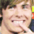 Pixelated close-up of a smiling person with light skin, brown hair, and visible teeth, set against a blurred background with dark and colorful elements.
