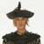 Pixelated figure wearing a wide-brimmed black hat with a tall central spike, dark clothing, and light skin tone against a plain background.