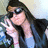 Pixelated image of a person with long dark hair wearing sunglasses and a dark jacket over a light blue shirt, holding a white object near their face.