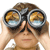 A person with brown hair holds black binoculars up to their eyes, showing a pixelated landscape reflection in the lenses.