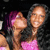 Two women in a close-up pose, one with purple hair and pink clothing, the other with long dark hair and a pink top, both smiling against a dark background.