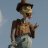 Pixelated figure in dark cap and green shirt holding brown object against light blue background.