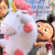 Animated cartoon character with dark hair in buns, wearing white polka-dot dress with pink accents, holding a pink object, against blurred background.