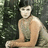 Pixelated image of a shirtless figure with light skin and dark hair, seated against a blurred green background.