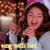 A person with long brown hair wearing a light blue top holds a black microphone, with blurred lights and a white object in the background and yellow text at the bottom.