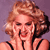 Pixelated image of a person with voluminous light brown hair styled in an updo, wearing a black-strapped top against a solid purple background.