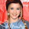 A person with light brown hair in a braid wears a blue top with white zigzag patterns against a red background with partial white text.
