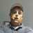 Pixelated static image of a person wearing a cap and dark clothing against a gray background.