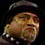 Pixelated close-up of a person wearing dark headwear and light-colored clothing against a black background.