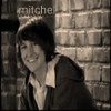 Black-and-white photo of a person with short dark hair smiling, wearing a dark jacket over a light collared shirt, standing against a brick wall with partial text 'mitche' visible above.