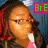 Pixelated image of a person with long dark hair and red highlights wearing glasses, pink top, and brown jacket against blue background with green text.