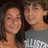 Two people in close proximity, one with long dark hair, the other in a gray shirt with white text, both smiling against a dark background.