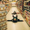 A person lies on a supermarket floor with arms and legs spread, holding a dark object above their head, surrounded by shelves of products.