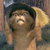 A pixelated figure with light brown hair wears a dark blue cap and holds a large yellow object near their face against a blurred background.