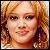 Static pixelated image of a person with light brown hair, fair skin, and dark eyes wearing small earrings.