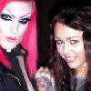 Two people pose closely together; one has bright pink hair and dark clothing, the other has long brown hair and a light-colored top.