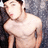 Pixelated image of a shirtless person with short dark hair, light skin tone, and visible facial features including eyes, nose, mouth, and ears against a blurred background.