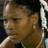 Pixelated static image of a person's face with brown skin tone and dark hair, wearing a white sleeveless top against a dark background.