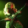 Static image of a person with long blonde hair playing an electric guitar on stage under green lighting.