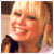 A pixelated image of a smiling person with blonde hair, wearing dark clothing and visible earrings.