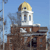 Pixelated static image of a building with a domed tower against a clear blue sky, featuring brown brickwork and white architectural details.