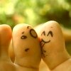 Two fingers with hand-drawn faces kissing against a blurred green background, one face showing surprise and the other smiling.