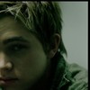 Close-up of a person's face with short dark hair, visible ear, and greenish clothing against a light background.