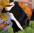 A pixelated cartoon character in a black suit and white shirt stands on green grass with a blue body of water and orange-brown background.