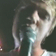A person with light brown hair sings into a teal microphone, wearing a dark shirt with green accents, against a dark background with faint pink lights.
