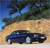 A static pixelated image shows a dark blue sedan on a sandy beach with trees and blue sky in the background.