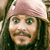 A pixelated close-up of a person wearing a light pink head covering with a small green accent, long dark hair, and a mustache, set against a blurred background.