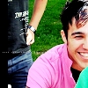 Two people stand outdoors on green grass; one wears a dark gray shirt with white text and blue jeans, the other a pink striped shirt.