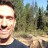 Pixelated static image of a person outdoors with trees and sky background, wearing dark clothing, mouth open in a smile or laugh.