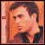Pixelated portrait of a person with dark hair and light skin, wearing a dark collared shirt against an orange-toned background.