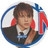 Pixelated circular icon shows a person with dark hair wearing a dark jacket over a white shirt, holding a red and blue object near their face.