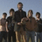 A pixelated group photo of six people in dark clothing against a light sky, with red pixelated text overlaid on a black background.