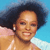 Pixelated close-up of a person with dark curly hair against a blue background, wearing a light-colored top and small earrings.