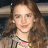 Pixelated static image of a person with long brown hair wearing a light-colored top and dark jacket, set against blurred background elements.
