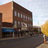 Pixelated static image of a multi-story building with brown and orange facades, blue awning, and street-level storefronts under a light sky.